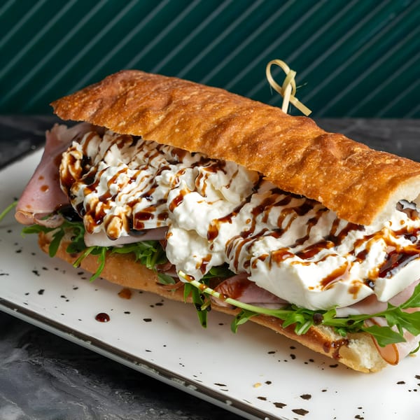 A SNDWCH baguette filled with ham, arugula, burrata cheese, and balsamic glaze on a white plate.
