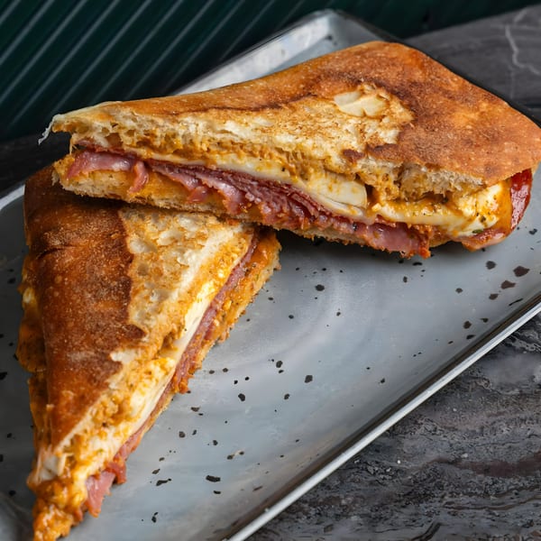 A toasted SNDWCH sandwich, cut in half, revealing layers of meat, cheese, and sauce on a gray plate.
