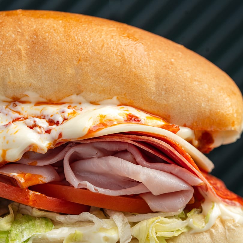 Close-up of a sub sandwich with melted cheese, pepperoni, ham, tomatoes, and lettuce.