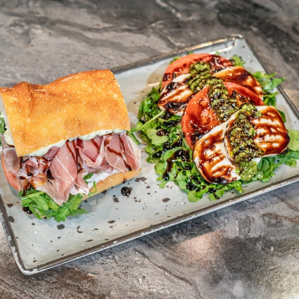 A SNDWCH prosciutto sandwich with a side of Caprese salad on a rectangular plate.