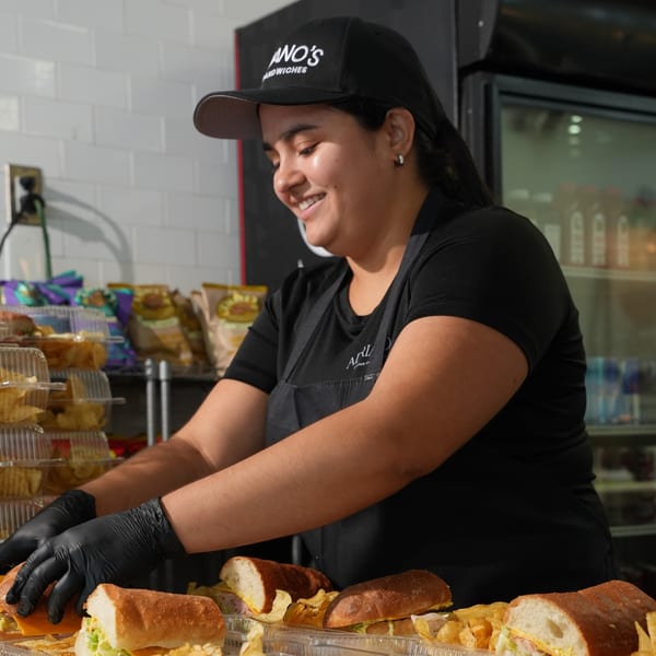 A smiling woman in a black cap and apron prepares sandwiches with chips in plastic containers.