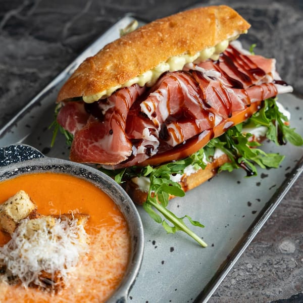 SNDWCH prosciutto sandwich with arugula and balsamic glaze, next to a bowl of tomato soup.