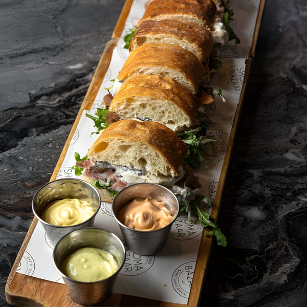 Sliced SNDWCH baguette sandwiches with meat, cheese, and arugula, served with three dipping sauces.