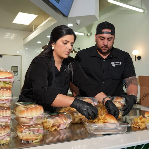 Two Adriano's employees in black shirts and gloves prepare sandwiches and chips for catering orders.