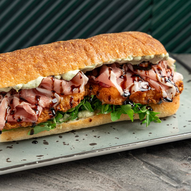 Close-up of a baguette sandwich filled with fried chicken, prosciutto, arugula, and a creamy sauce.