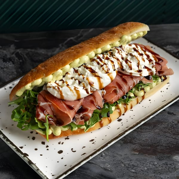 SNDWCH baguette with prosciutto, burrata, arugula, avocado cream, and balsamic glaze on a white plate.