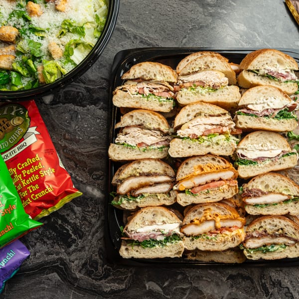 Catering tray of assorted sandwiches, a bowl of Caesar salad, and bags of chips on a dark counter.