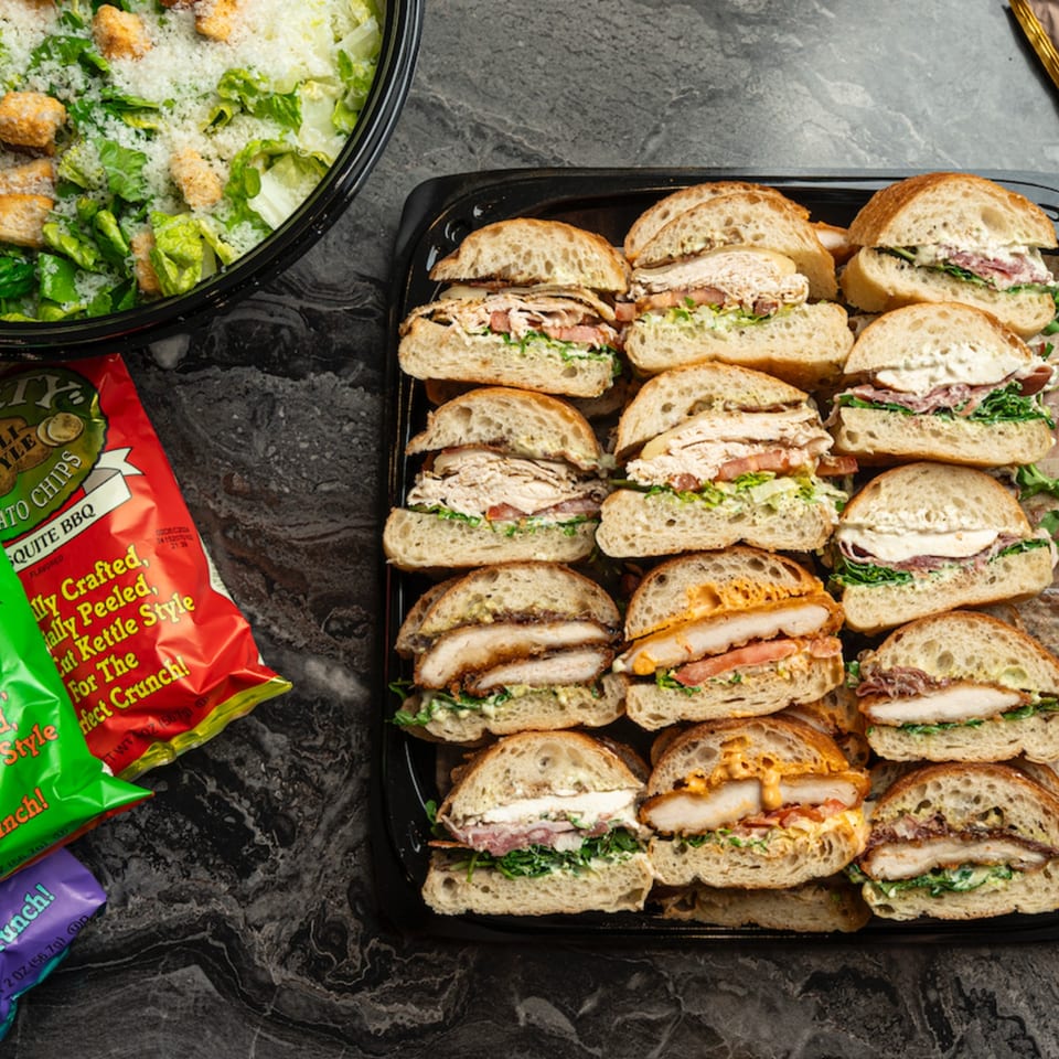 Catering tray of assorted sandwiches, a bowl of Caesar salad, and bags of chips on a dark counter.