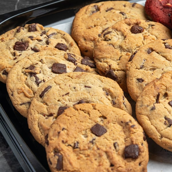 Close-up of a tray filled with golden-brown chocolate chip cookies, with one red velvet cookie visible.