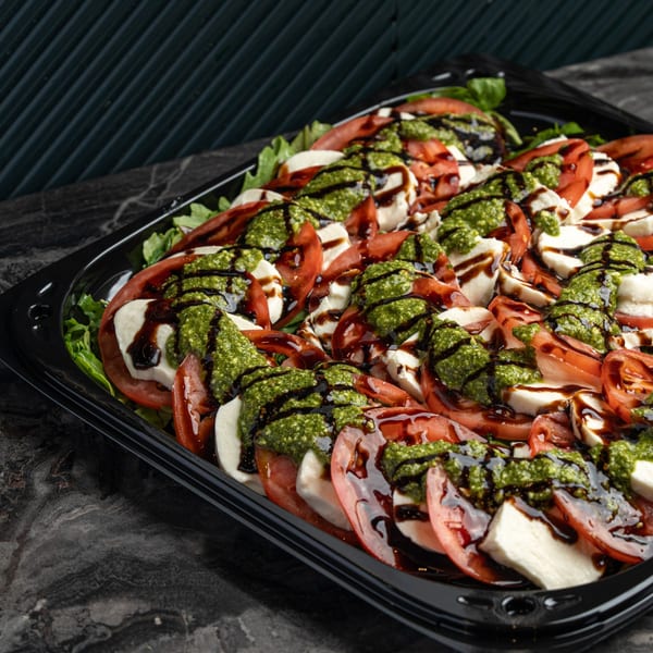 Caprese salad platter with sliced tomatoes, mozzarella, pesto, and balsamic glaze on a black tray.