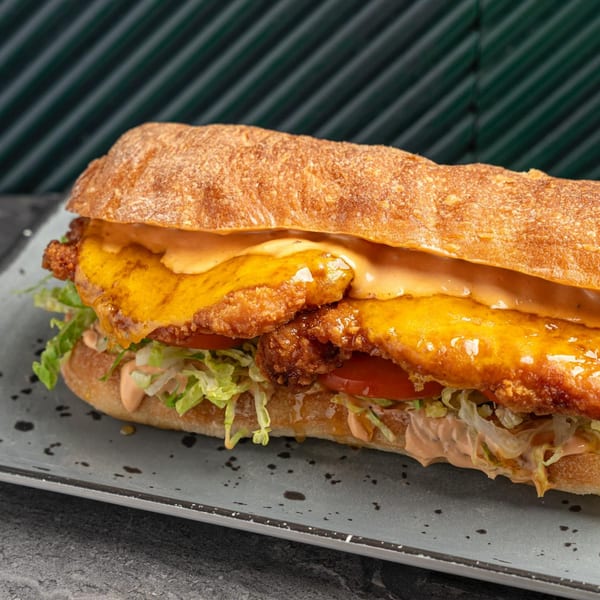 SNDWCH fried chicken sandwich with melted cheese, lettuce, tomatoes, and sauce on a baguette.