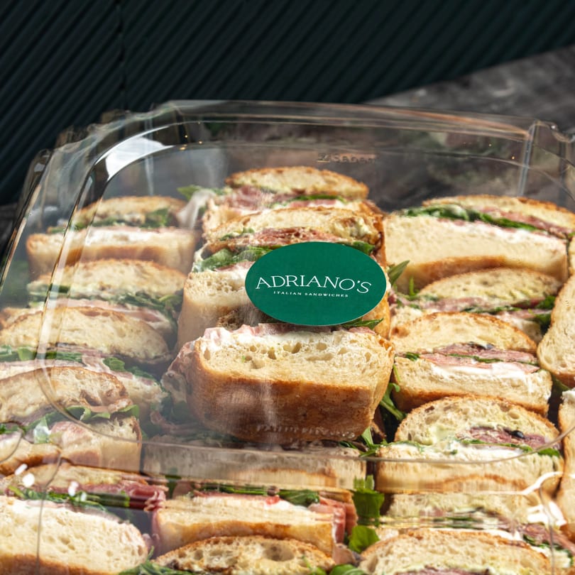 Adriano's Italian Sandwiches catering tray filled with sliced sandwiches, prosciutto, arugula, and cream cheese.