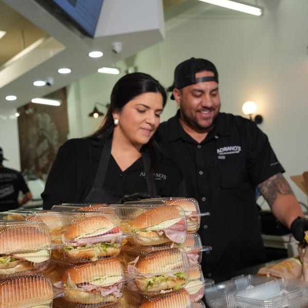 Two Adriano's employees smiling while preparing numerous pre-packaged sandwiches for customers.